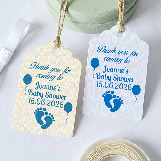 Two personalised baby shower gift tags with blue text and baby footprint/balloon graphics, shown on a white background with ribbon and tape props.
