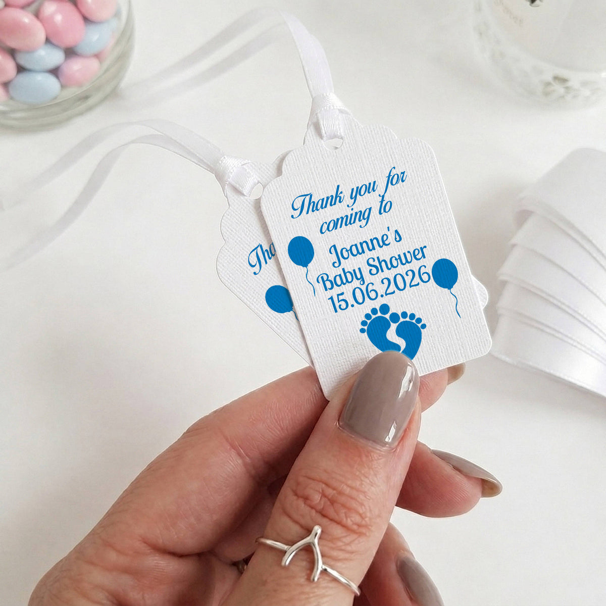 A hand holding two white personalised baby shower favour tags with blue text and baby footprints, set against a soft white background with blurred decorative candies and ribbon.