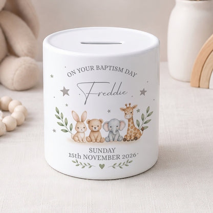 A white ceramic baby baptism money box with a personalised print featuring cartoon animals and the text 'ON YOUR BAPTISM DAY Freddie SUNDAY 15th NOVEMBER 2026'.