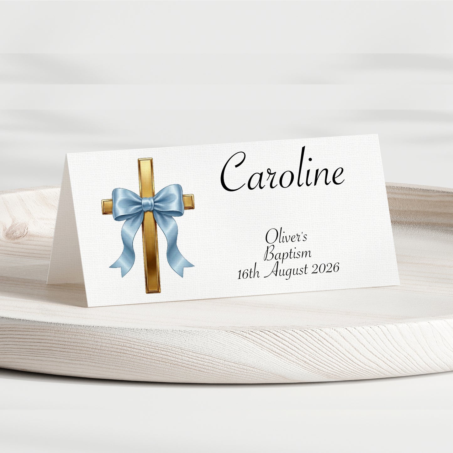 A personalised baptism place card standing on a white wooden surface. The card is white with a printed gold cross, a light blue bow illustration, and the name 'Caroline' along with the text 'Oliver's Baptism 16th August 2026' in black script.