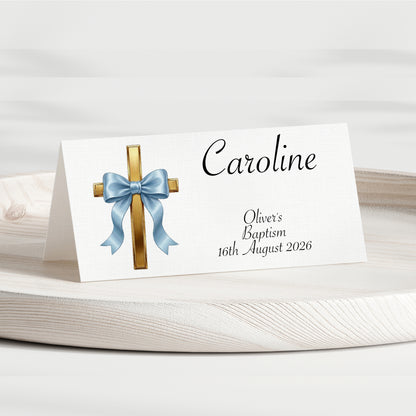 A personalised baptism place card standing on a white wooden surface. The card is white with a printed gold cross, a light blue bow illustration, and the name 'Caroline' along with the text 'Oliver's Baptism 16th August 2026' in black script.