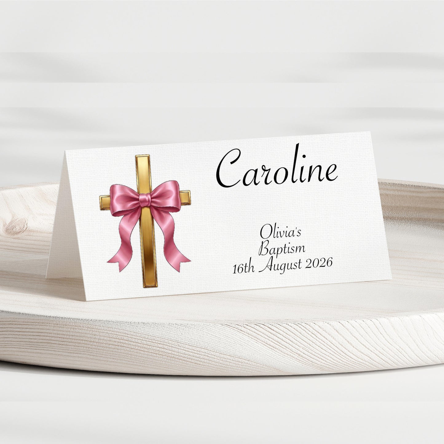 A personalised baptism place card on a white folding stand, featuring a gold cross with a pink satin bow illustration and the name Caroline, with custom text for Olivia's Baptism dated 16th August 2026.