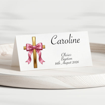 A personalised baptism place card on a white folding stand, featuring a gold cross with a pink satin bow illustration and the name Caroline, with custom text for Olivia's Baptism dated 16th August 2026.