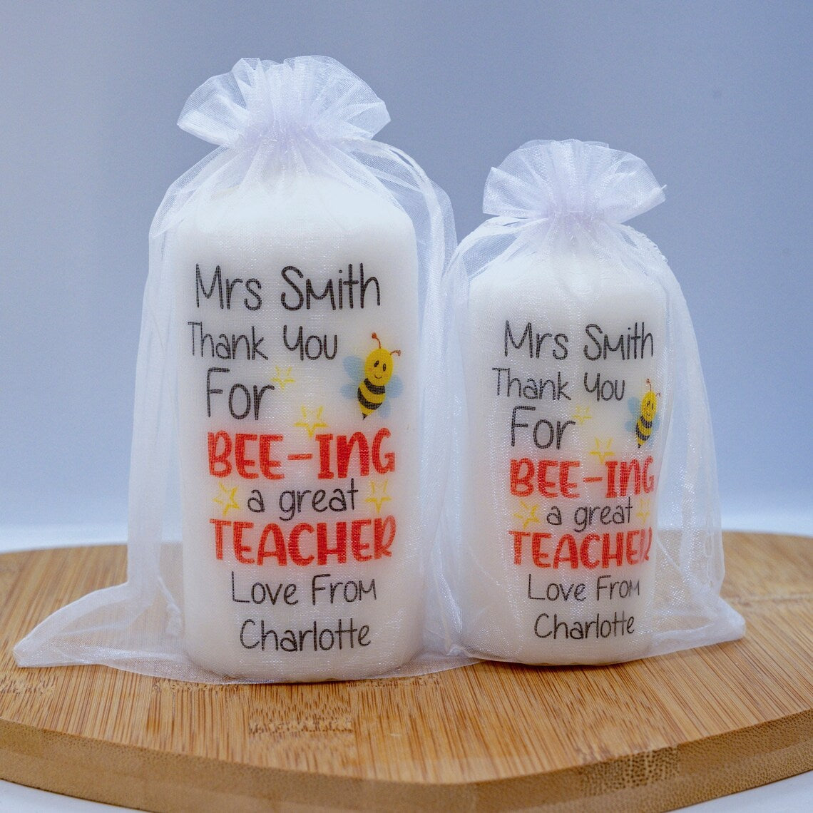 Two personalised candles with bee illustrations and the message 'Thank You For Bee-ing A Great Teacher', with a space for personalisation at the bottom, displayed in clear packaging on a wooden board.