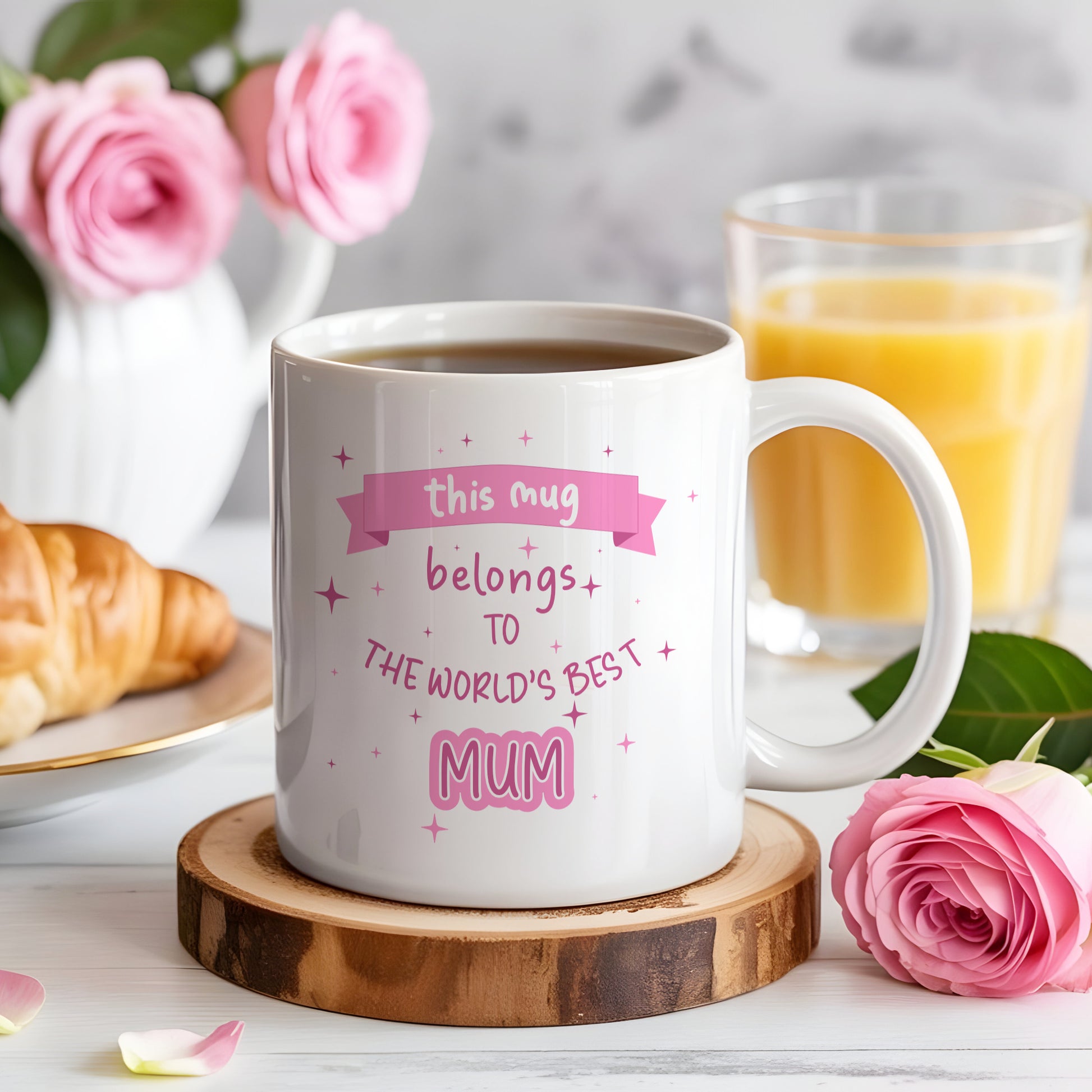 White mug with 'world's best mum' text on a wooden coaster with coffee, croissant, and flowers.