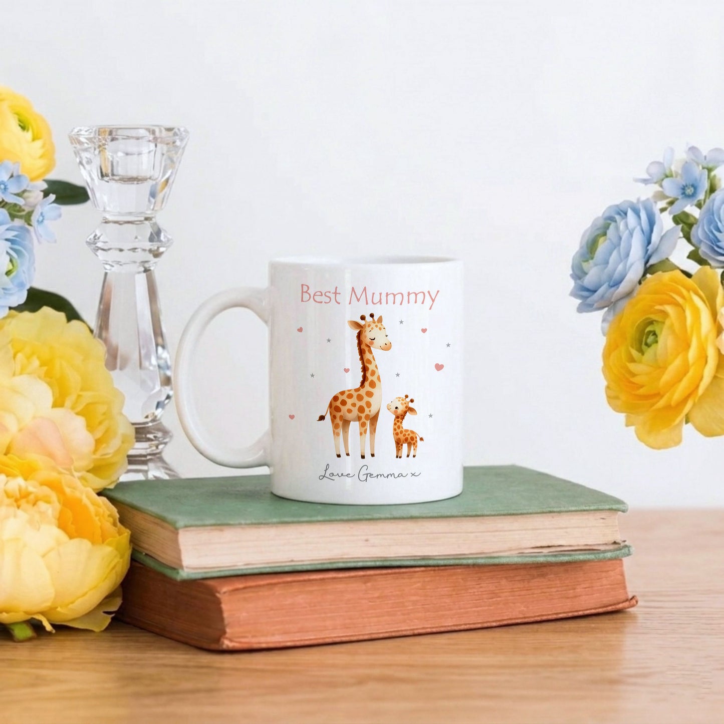 A white ceramic mug with a cute giraffe illustration and the text 'Best Mummy' and 'Love, Grammar' sits on a stack of vintage books on a wooden table, styled with yellow and blue flowers and a glass candlestick.