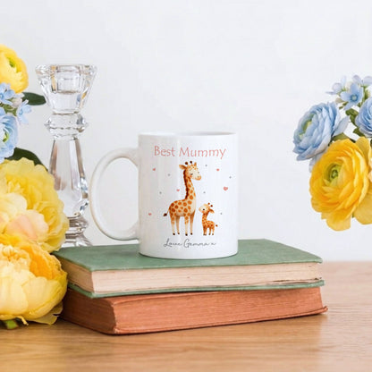 A white ceramic mug with a cute giraffe illustration and the text 'Best Mummy' and 'Love, Grammar' sits on a stack of vintage books on a wooden table, styled with yellow and blue flowers and a glass candlestick.