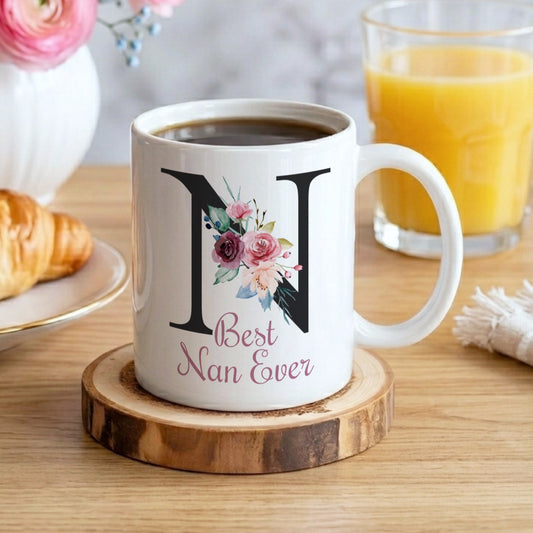 A white ceramic coffee mug with a large black letter 'N' and floral design, printed with 'Best Nan Ever' in pink script, sitting on a wooden coaster on a wooden table. A croissant is on a plate to the left, a glass of orange juice is to the right, and a vase of pink flowers is in the background.