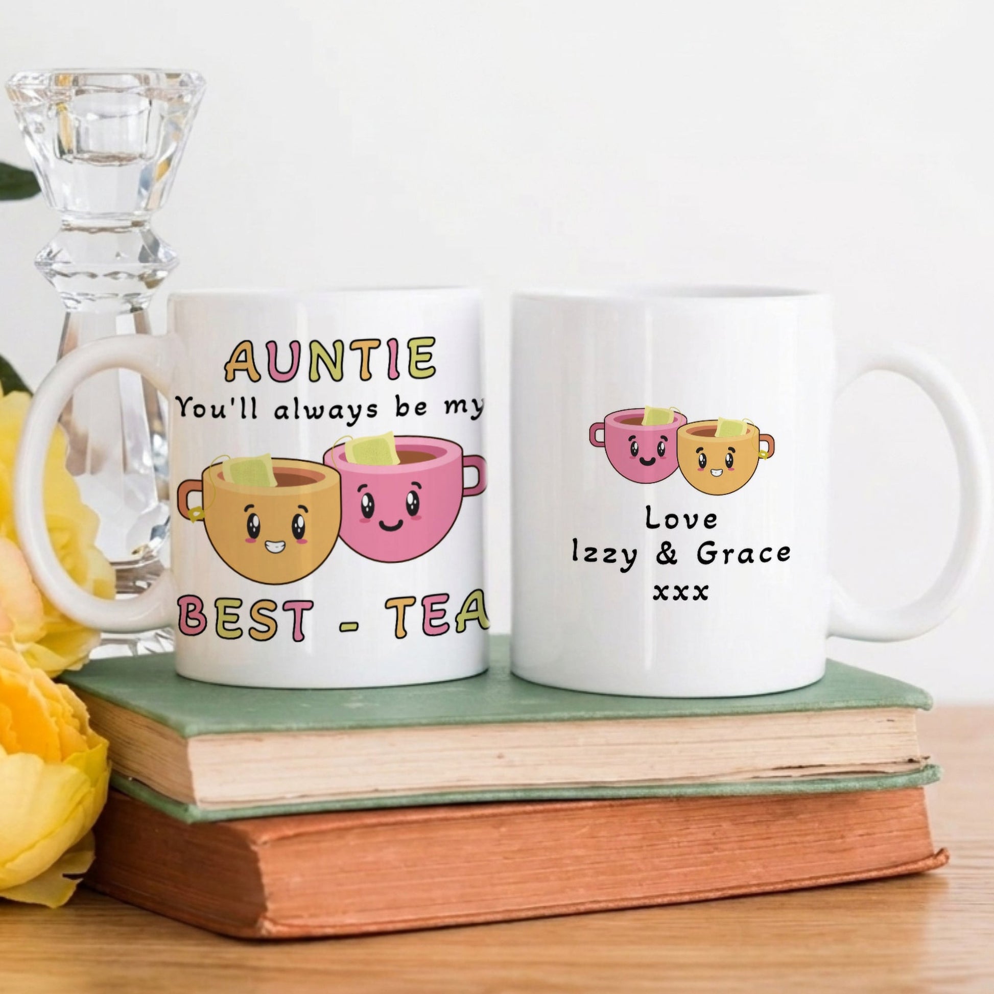 Two white ceramic mugs with cute cartoon tea cup graphics and personalised text, displayed on stacked books with yellow flowers and a glass candlestick holder in the background.