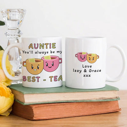 Two white ceramic mugs with cute cartoon tea cup graphics and personalised text, displayed on stacked books with yellow flowers and a glass candlestick holder in the background.