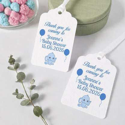 Two white baby shower favour tags with blue text and illustrations of a blue elephant and balloons, styled on a light grey surface with a green round box, a glass bowl of pastel candies, and a sprig of eucalyptus.