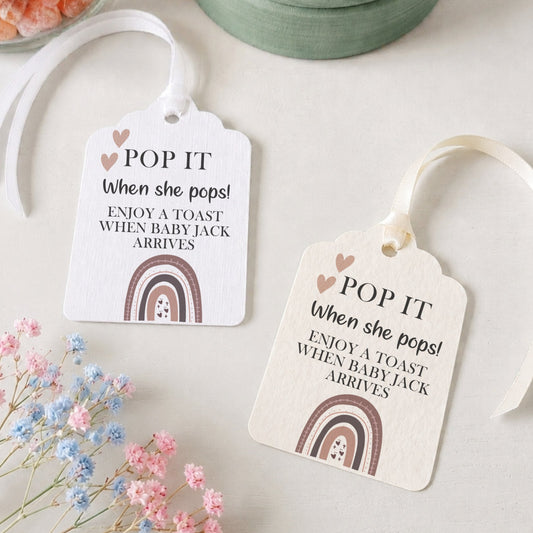 Two white paper gift tags with white ribbons, printed with the phrase 'POP IT When she pops! ENJOY A TOAST WHEN BABY JACK ARRIVES' and a small rainbow illustration, styled on a light surface with dried flowers, a jar of orange peels, and a round green box.