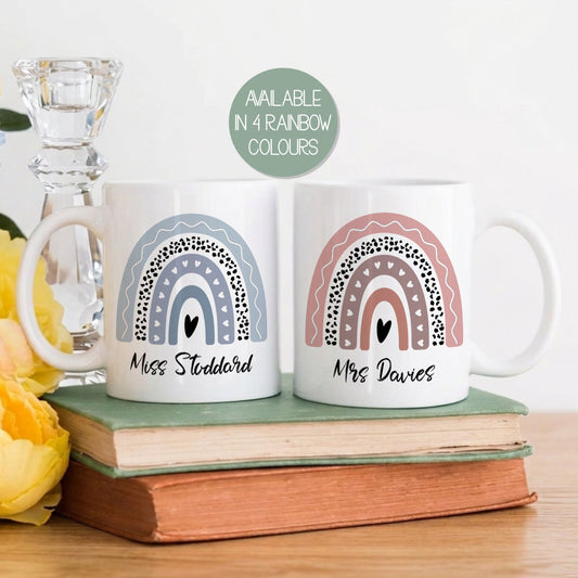 Two mugs with rainbow designs and personalized names on a wooden surface with flowers and books.