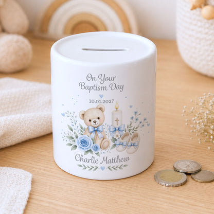 A personalised white ceramic baptism money box on a wooden tabletop, decorated with a watercolour teddy bear, flowers, baby booties, and the text 'On Your Baptism Day' and a custom name.