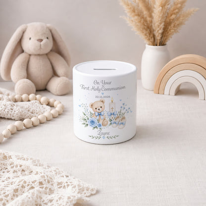 A personalised white ceramic First Holy Communion money box with a teddy bear illustration and the name 'Zayne' printed on it, styled on a light fabric surface with a plush bunny, wooden beads, a vase with dried grass, and a wooden rainbow in the background.