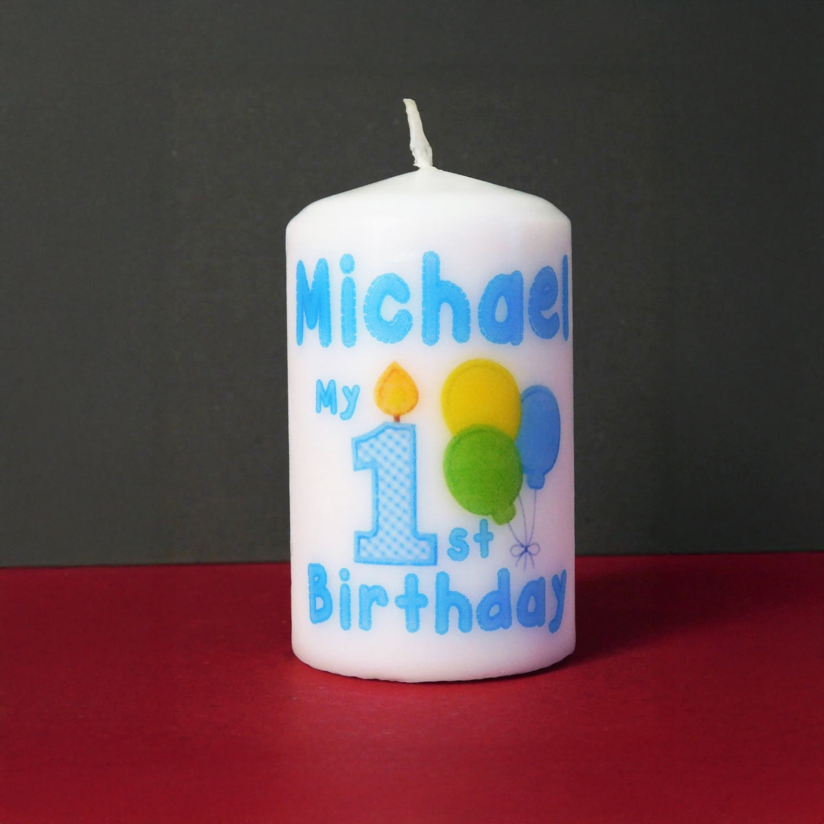 A white pillar candle with blue text reading 'Michael My 1st Birthday' and colourful balloon graphics, set against a dark grey background and red surface.