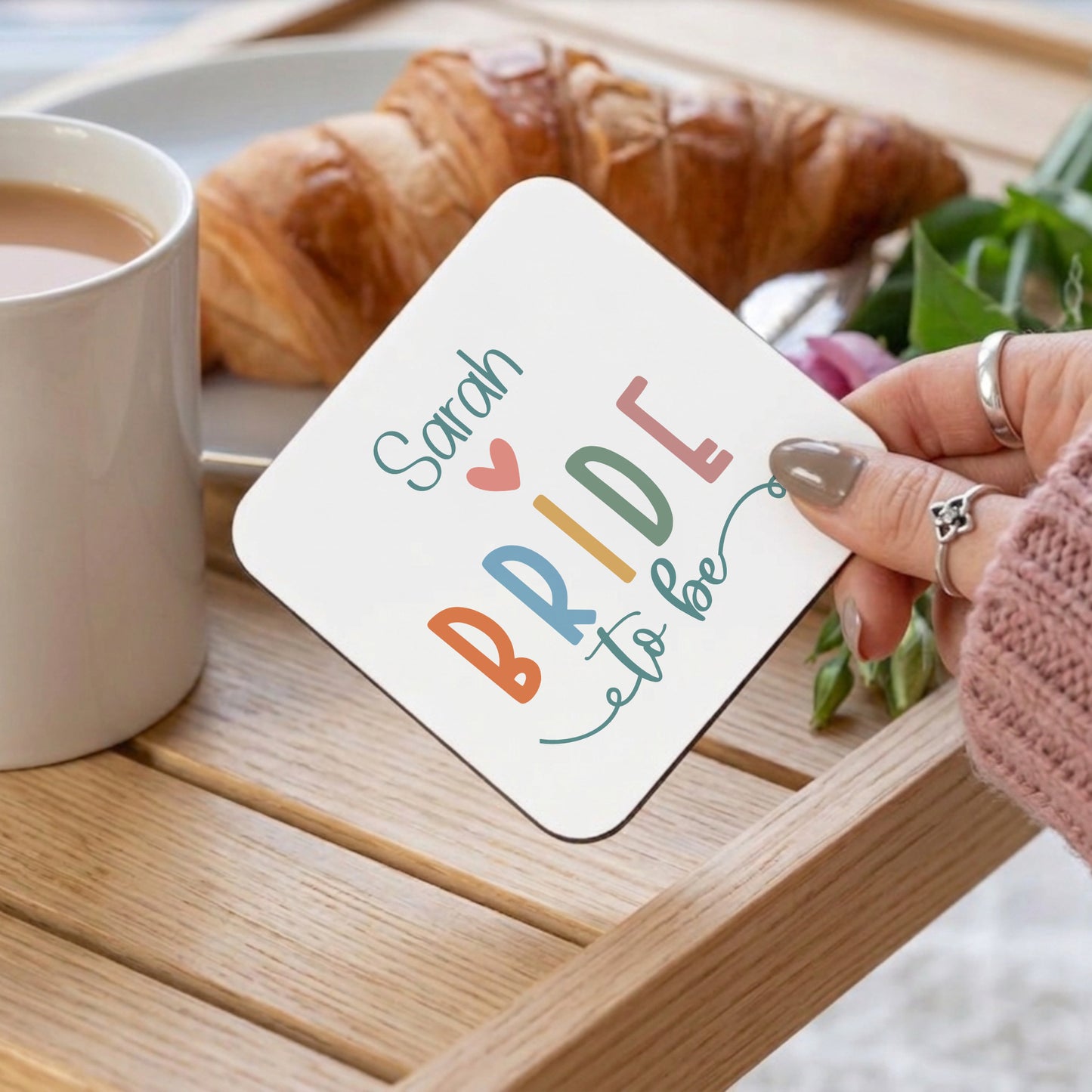 Coaster with 'Sarah Bride to be' text held by a hand on a table with coffee and croissant.