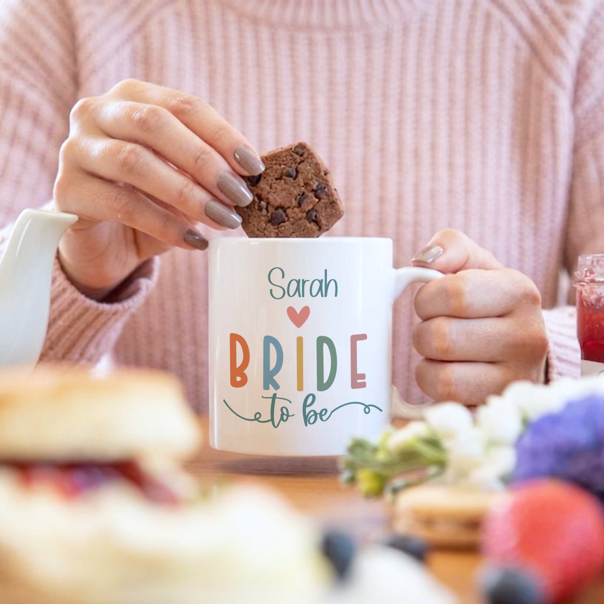 Person holding a mug labeled 'Sarah Bride to be' with a cookie, blurred background