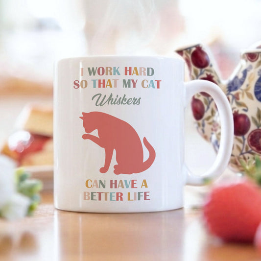 A white ceramic mug with a red cat silhouette and the text 'I WORK HARD SO THAT MY CAT Whiskers CAN HAVE A BETTER LIFE' sits on a wooden table in a bright kitchen setting, with a patterned teapot, strawberries, and flowers blurred in the background.