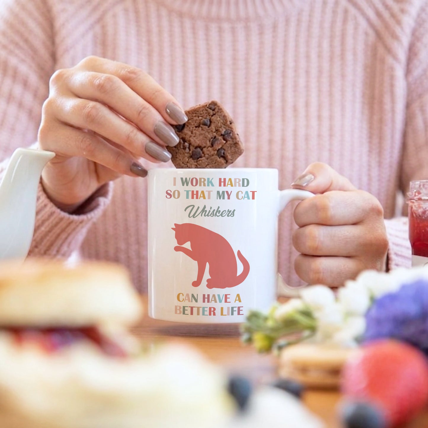 A person in a pink ribbed sweater holds a white ceramic mug with a red cat silhouette and the text 'I WORK HARD SO THAT MY CAT Whiskers CAN HAVE A BETTER LIFE' while placing a chocolate chip cookie into the mug. The scene is set on a table with breakfast items in a blurred foreground and background.