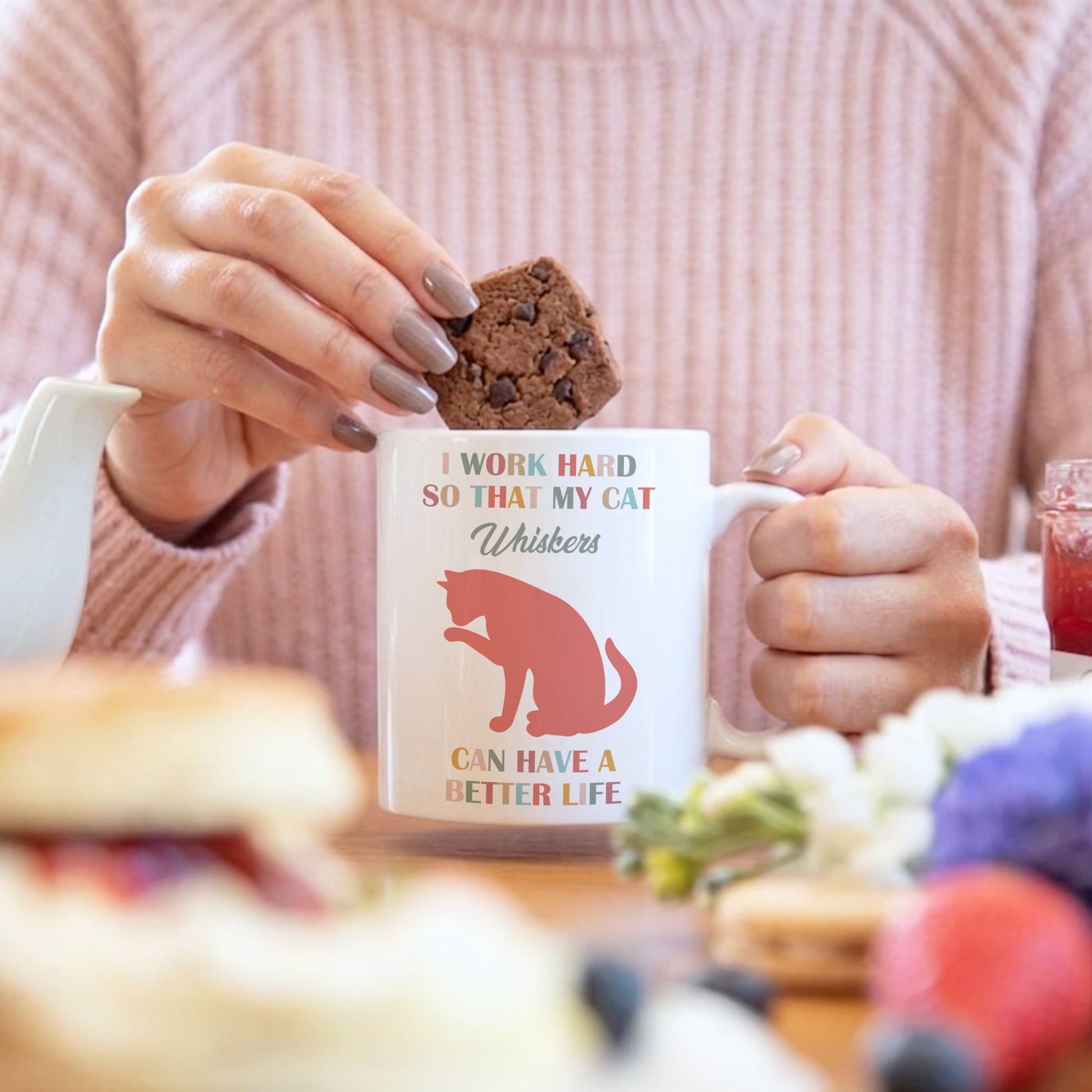A person in a pink ribbed sweater holds a white ceramic mug with a red cat silhouette and the text 'I WORK HARD SO THAT MY CAT Whiskers CAN HAVE A BETTER LIFE' while placing a chocolate chip cookie into the mug. The scene is set on a table with breakfast items in a blurred foreground and background.