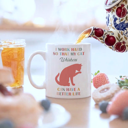 A white ceramic mug with a colorful printed slogan and cat illustration sits on a table in a breakfast setting, with a patterned teapot pouring tea into it and strawberries, donuts, and a jar of iced tea nearby.