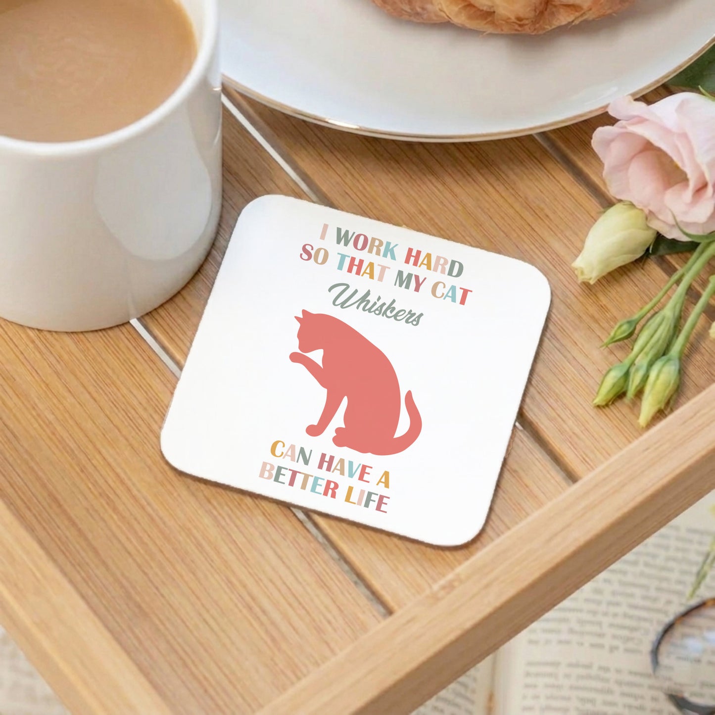A square white coaster with rounded corners sits on a wooden tray. The coaster has a red cat silhouette and the text 'I WORK HARD SO THAT MY CAT Whiskers CAN HAVE A BETTER LIFE' in multicoloured lettering.