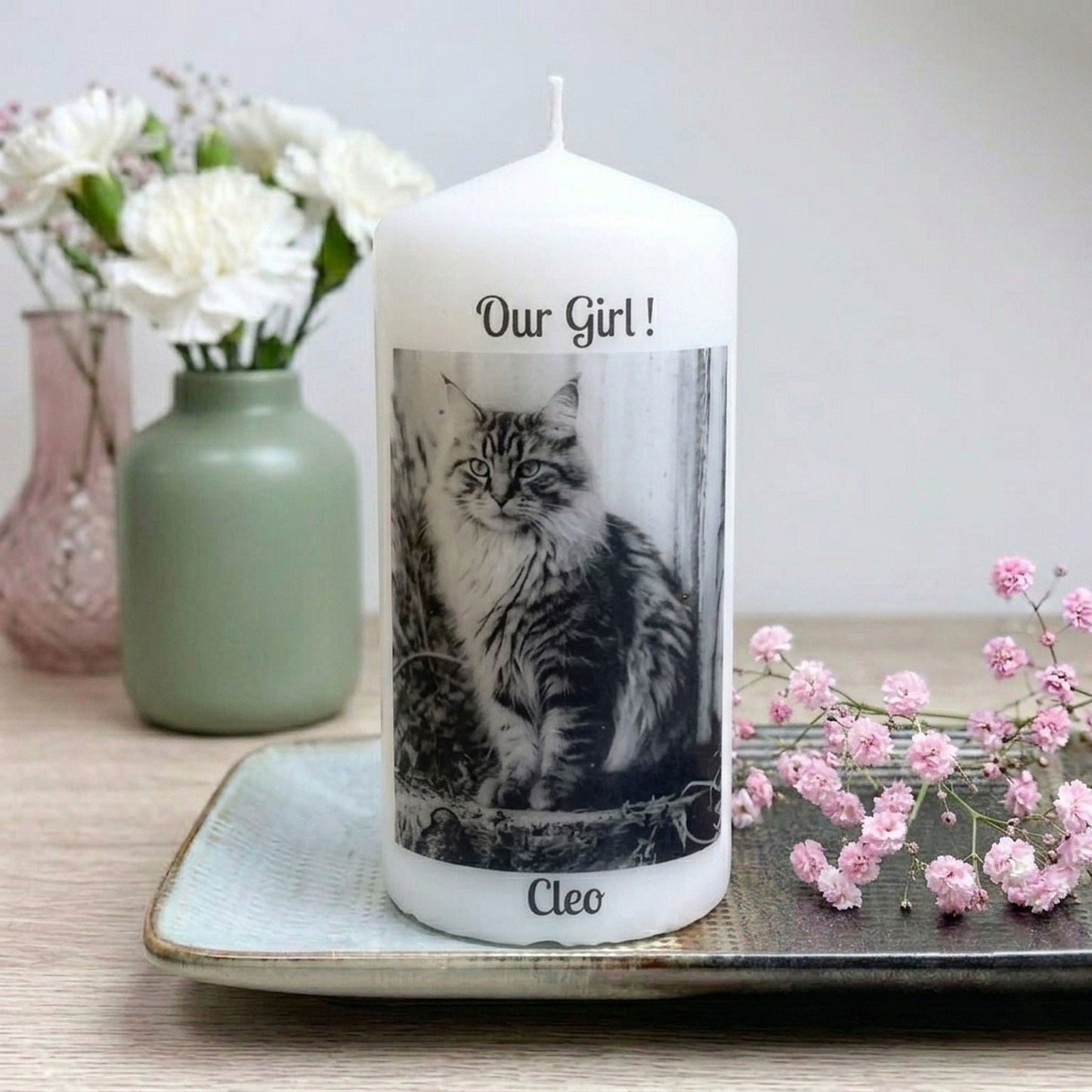 A white taper candle with a custom photo print of a cat labeled 'Our Girl!' and the name 'Cleo', displayed on a tray with decorative flowers in the background.
