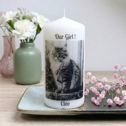 A white taper candle with a custom photo print of a cat labeled 'Our Girl!' and the name 'Cleo', displayed on a tray with decorative flowers in the background.