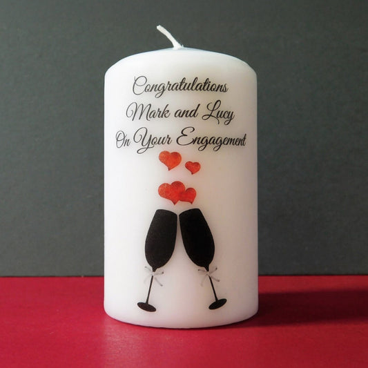 White candle with engagement congratulations text and wine glass illustrations on a red surface.
