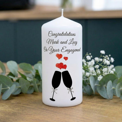 A white taper candle with a personalised engagement message and a champagne glass graphic, styled on a wooden surface with eucalyptus leaves and baby's breath flowers.