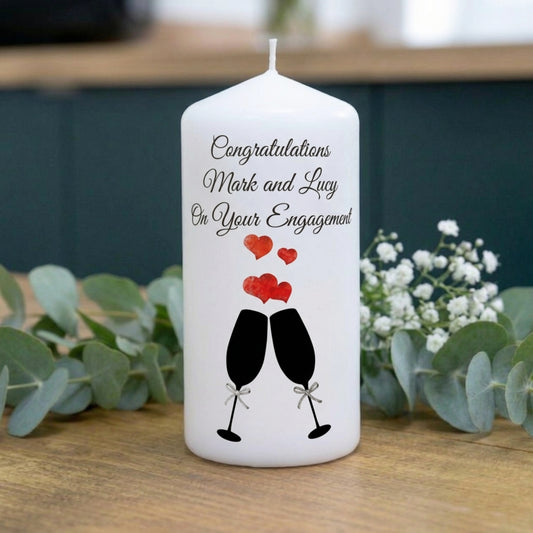 A white taper candle with a personalised engagement message and a champagne glass graphic, styled on a wooden surface with eucalyptus leaves and baby's breath flowers.