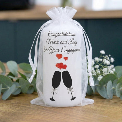 A white cylindrical bottle or jar wrapped in a sheer white organza drawstring bag, printed with the message 'Congratulations Mark and Lucy On Your Engagement' above a black-and-red clinking glasses and hearts graphic. The bag sits on a wooden surface with greenery and small white flowers in the background.