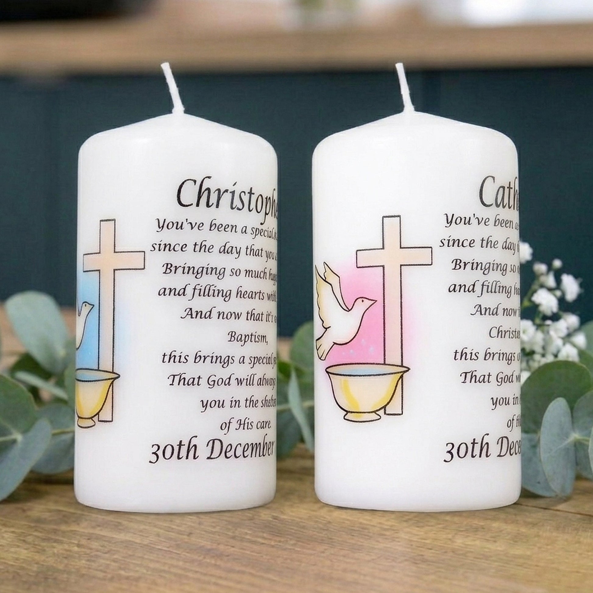 Two personalised white pillar candles with printed Christian memorial text and religious symbols, displayed on a wooden surface with greenery in the background.