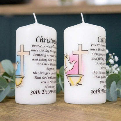 Two personalised white pillar candles with printed Christian memorial text and religious symbols, displayed on a wooden surface with greenery in the background.
