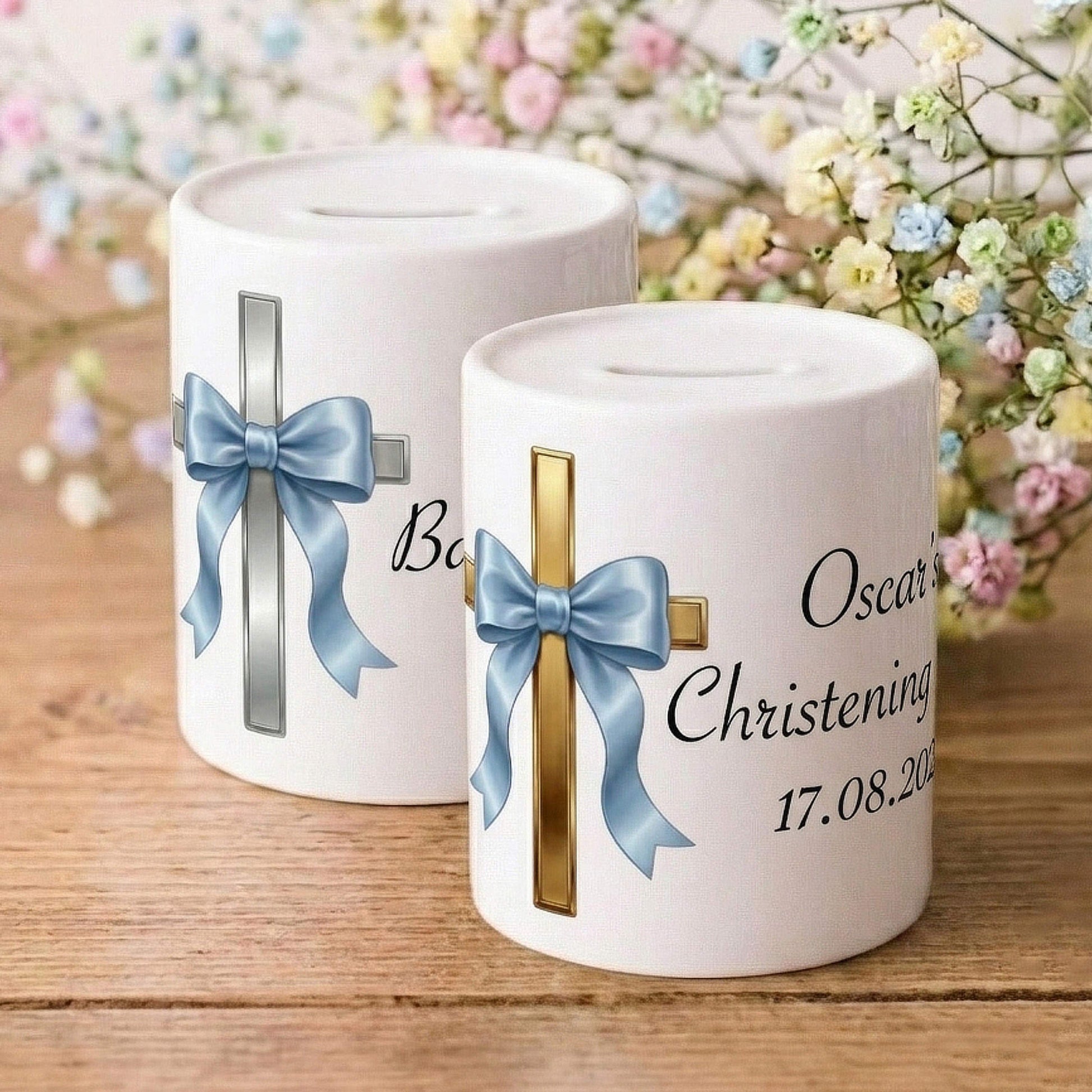 Two white ceramic Christening or Baptism money canisters on a wooden surface, decorated with blue and gold crosses and bows, with baby's breath flowers in the background.