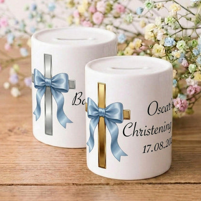 Two white ceramic Christening or Baptism money canisters on a wooden surface, decorated with blue and gold crosses and bows, with baby's breath flowers in the background.