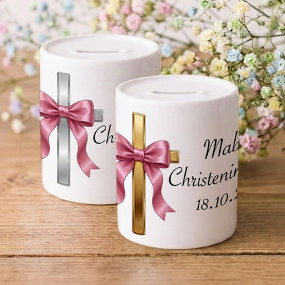 Two white ceramic baby christening money boxes on a wooden surface with baby's breath flowers in the background. One box features a silver cross with a pink bow and the text 'Chri.