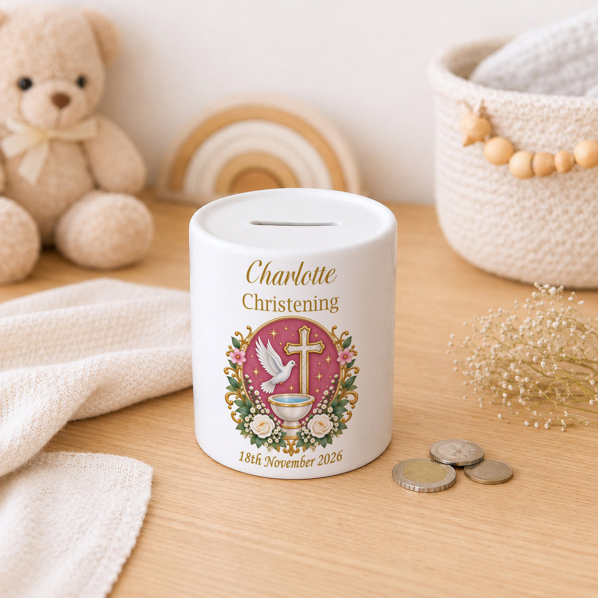 A white ceramic baby christening money box with a custom printed illustration of a dove, cross, and flowers, personalized with the name 'Charlotte' and date '18th November 2026'. It sits on a wooden table surrounded by a teddy bear, knit blankets, and coins.