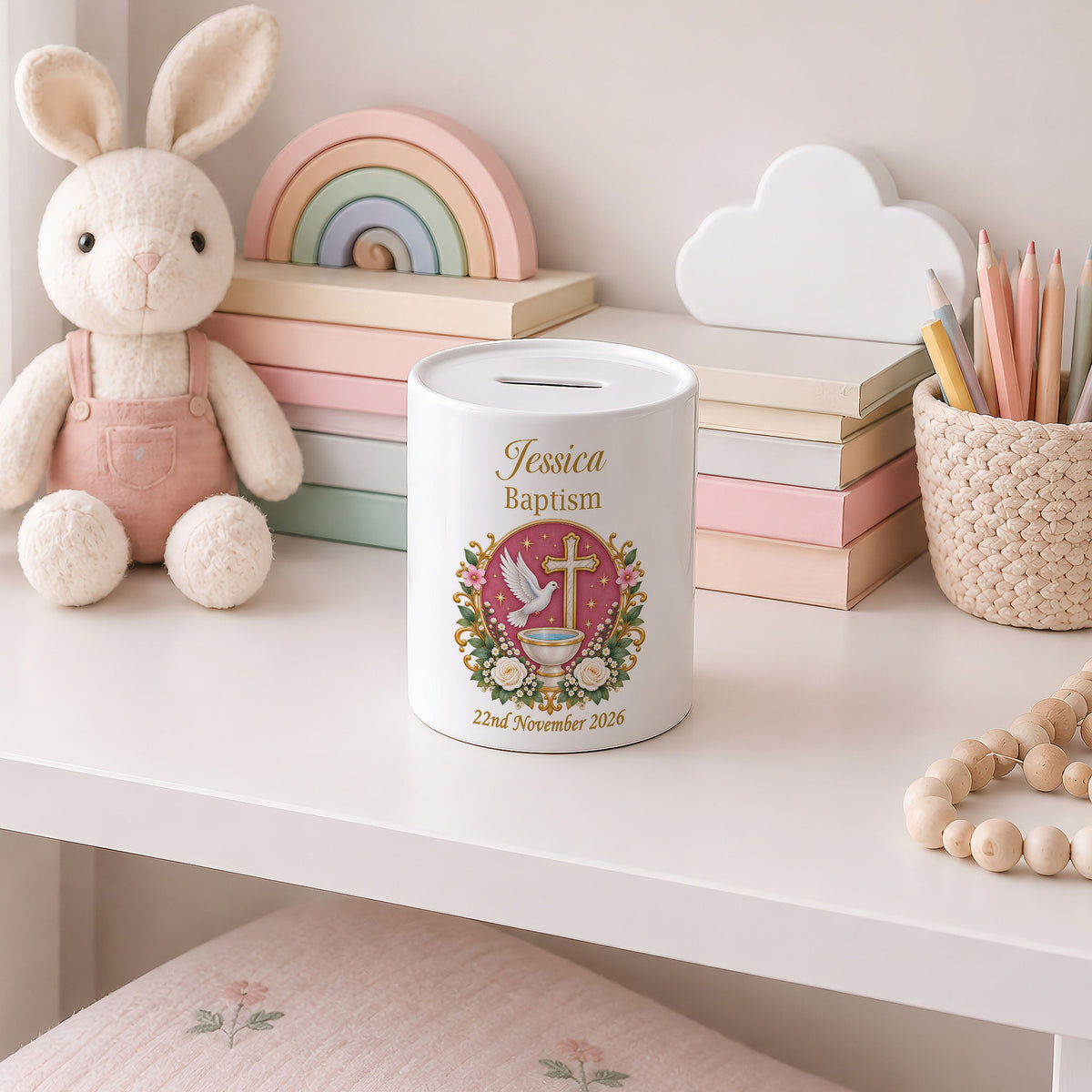 A white cylindrical baptism money box with a slot on top, personalised with the name 'Jessica Baptism' and a colourful cross design, displayed on a white shelf next to a plush bunny, pastel books, and stationery.