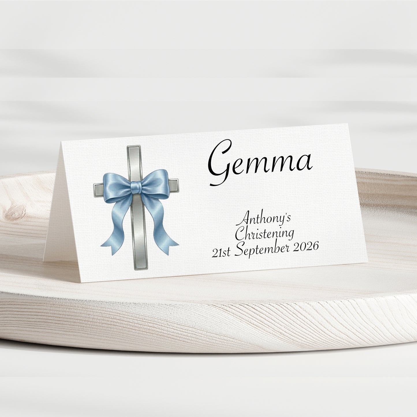 A personalised place card for a christening, featuring a white background with a silver cross decorated with a light blue satin bow and the name 'Gemma' in black script. The card is displayed on a round wooden tray against a soft, light gray background.