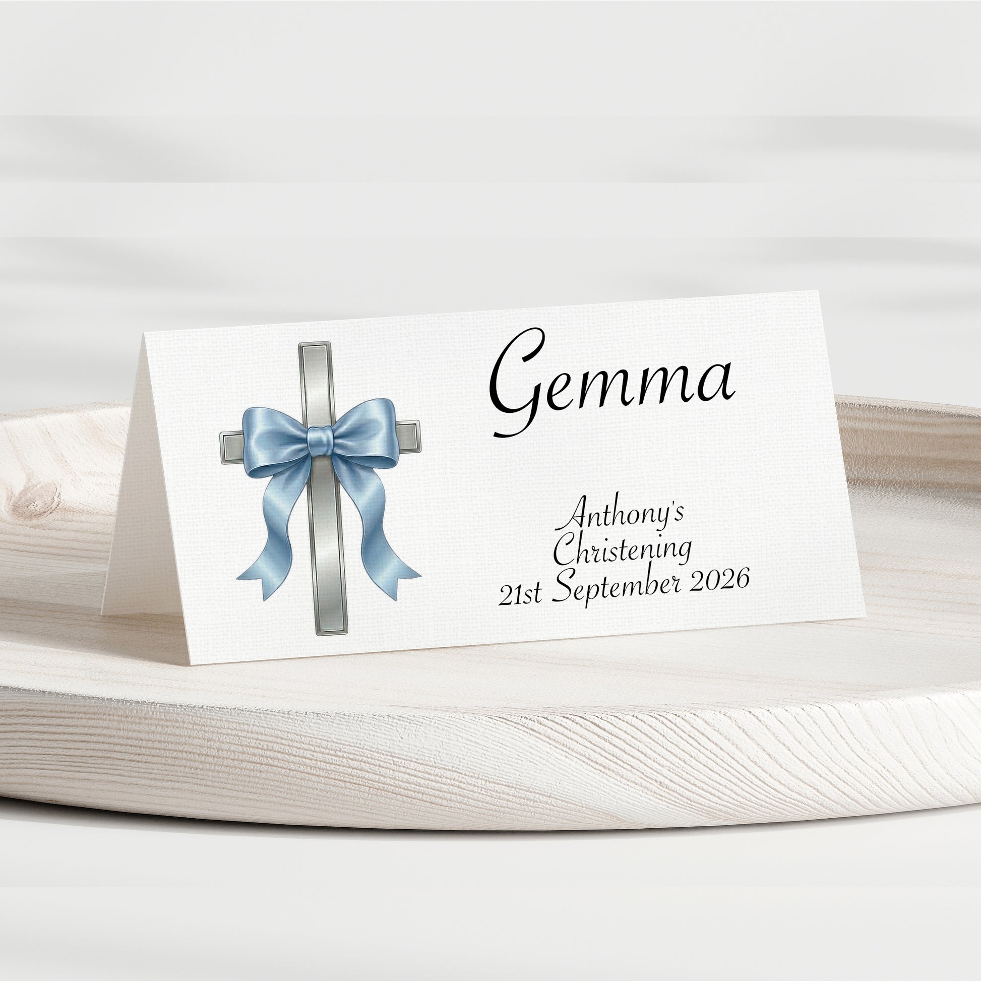 A personalised place card for a christening, featuring a white background with a silver cross decorated with a light blue satin bow and the name 'Gemma' in black script. The card is displayed on a round wooden tray against a soft, light gray background.