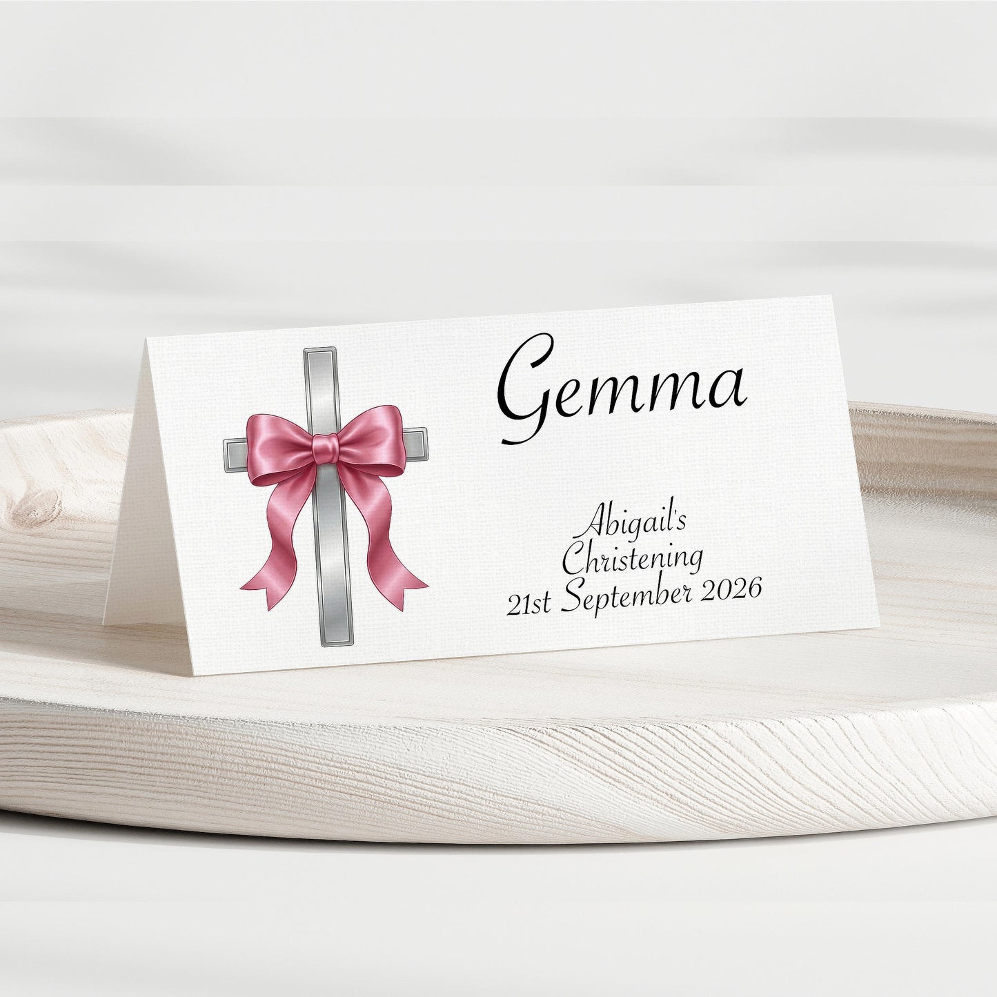 A personalised place card for a christening, featuring a white background with a silver cross and pink bow graphic, and the name 'Gemma' along with the event details printed in black script.