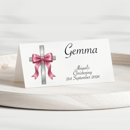 A personalised place card for a christening, featuring a white background with a silver cross and pink bow graphic, and the name 'Gemma' along with the event details printed in black script.