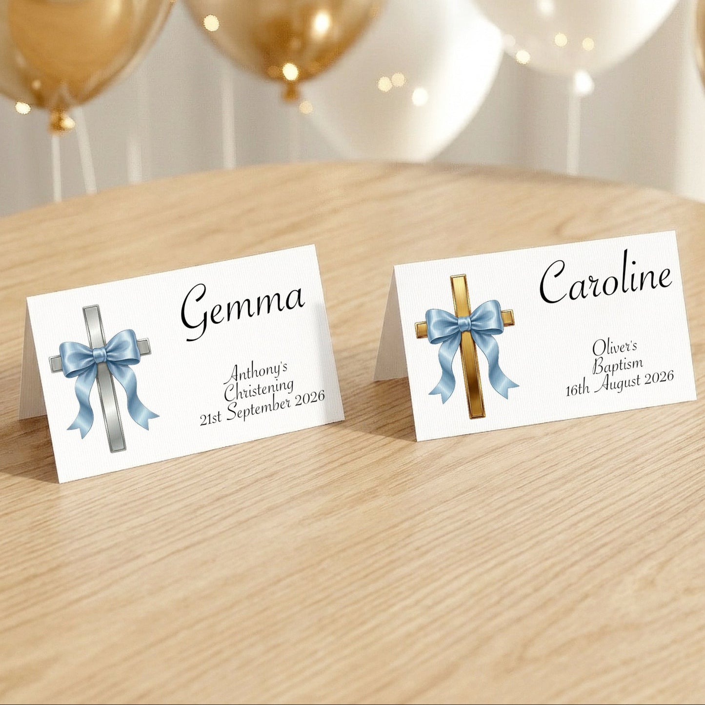 Two personalised baptism place cards standing on a light wood table, with gold and white balloons blurred in the background. One card has a silver cross with a blue ribbon and the name Gemma, and the other has a gold cross with a blue ribbon and the name Caroline.