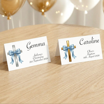 Two personalised baptism place cards standing on a light wood table, with gold and white balloons blurred in the background. One card has a silver cross with a blue ribbon and the name Gemma, and the other has a gold cross with a blue ribbon and the name Caroline.