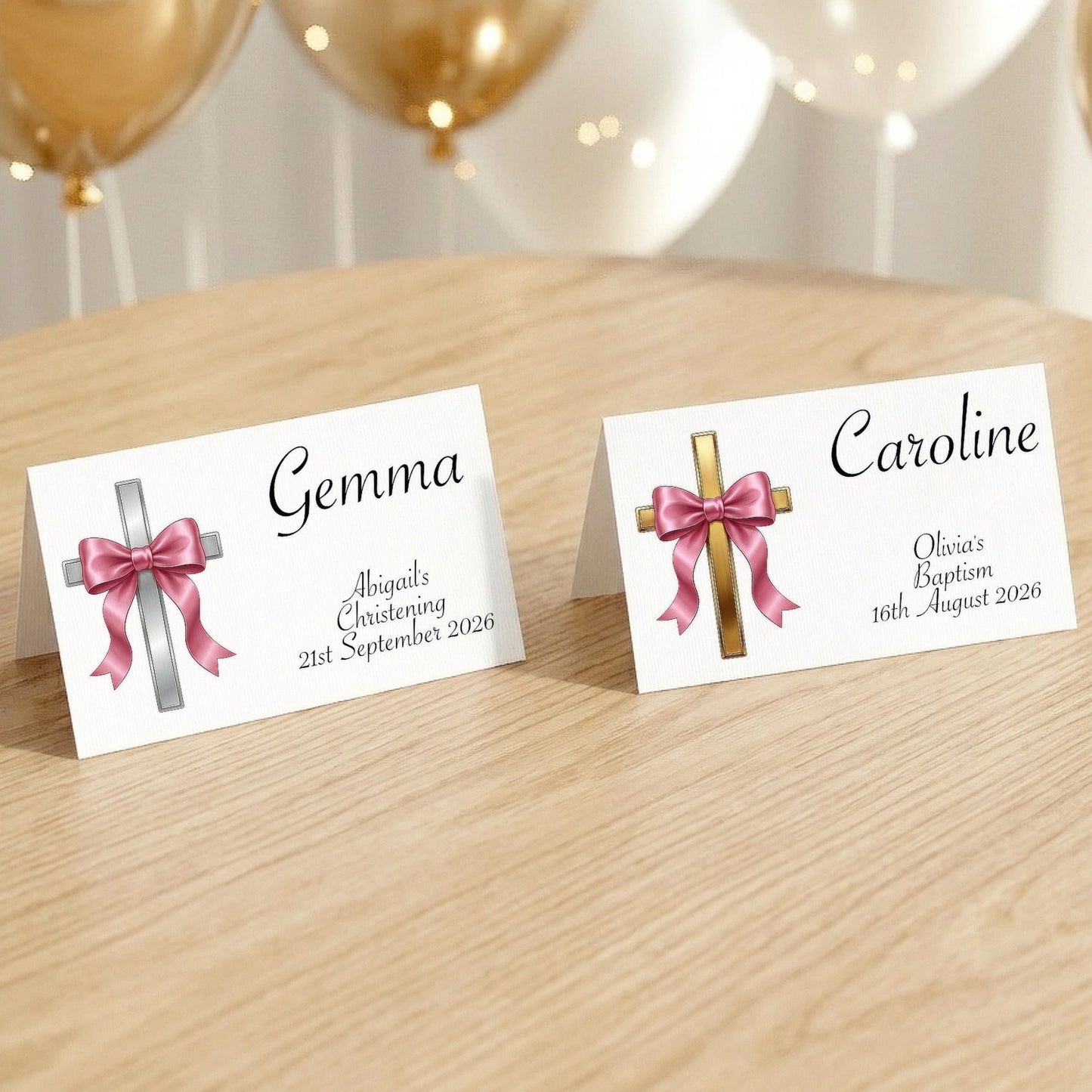 Two personalised baptism place cards standing on a light wood table, with gold and white balloons blurred in the background. One card has a silver cross with a pink bow and the name Gemma, and the other has a gold cross with a pink bow and the name Caroline.