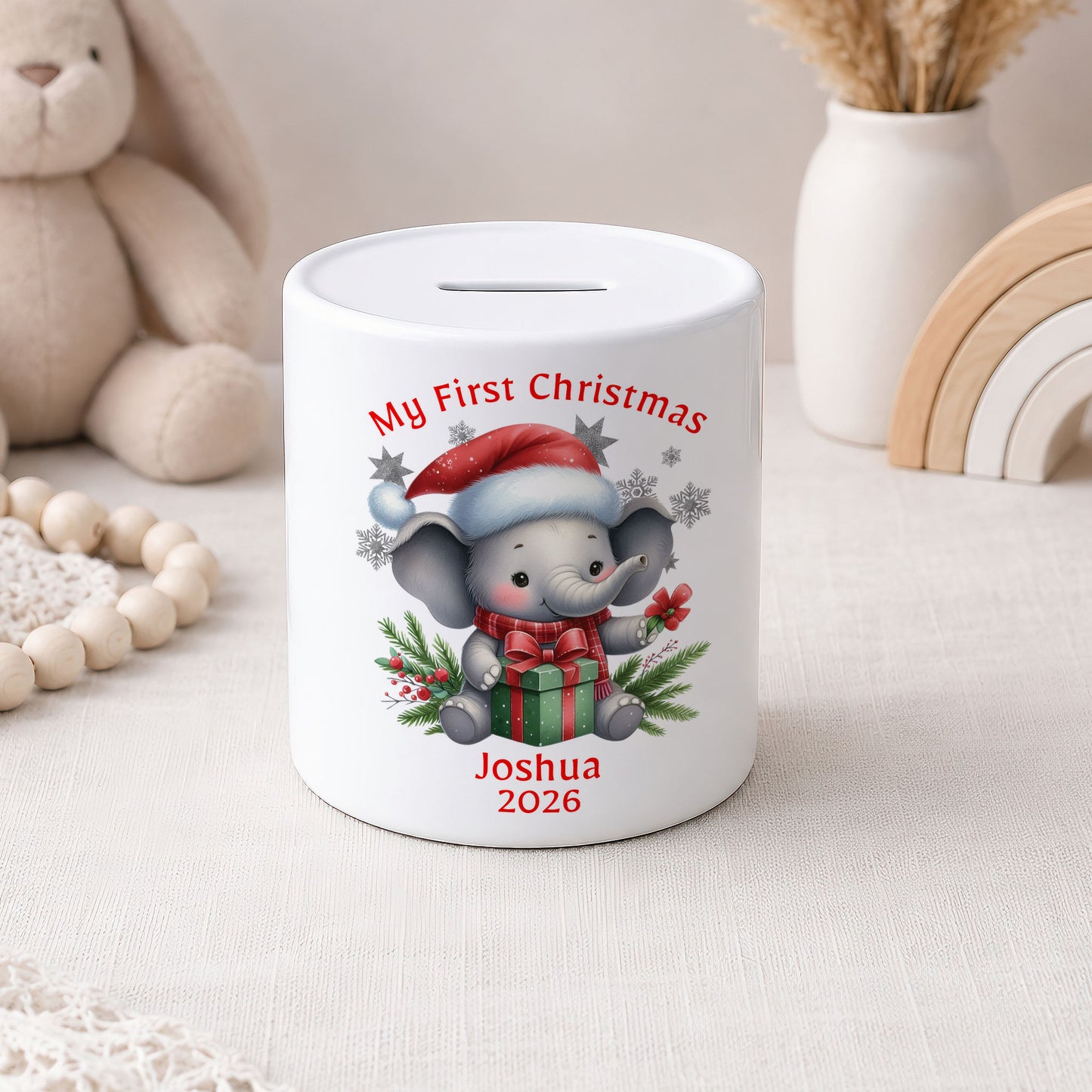 A white ceramic pig bank with a custom cartoon elephant illustration and the text 'My First Christmas Joshua 2026'. The bank is styled on a light fabric surface with a wooden bead garland, a plush bunny, and a white vase in the background.