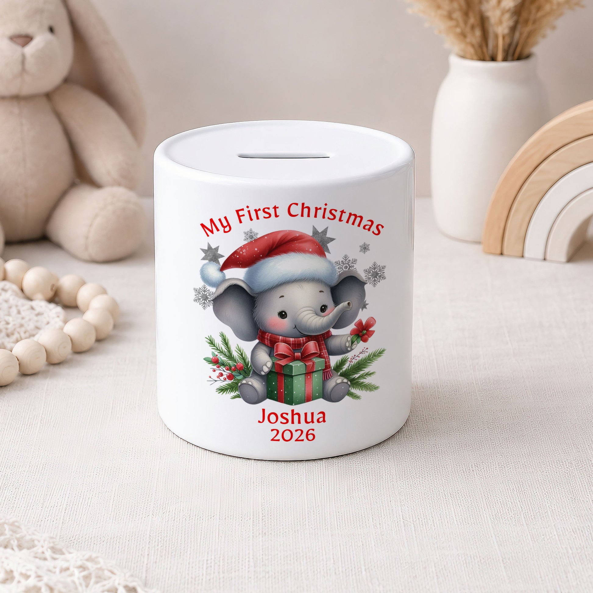 A white ceramic pig bank with a custom cartoon elephant illustration and the text 'My First Christmas Joshua 2026'. The bank is styled on a light fabric surface with a wooden bead garland, a plush bunny, and a white vase in the background.