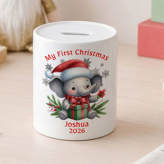 A white cylindrical pig bank decorated with a cartoon elephant wearing a Santa hat and scarf, holding a gift box, with the text 'My First Christmas' and 'Joshua 2026'. The pig bank is set on a light wooden table with coins nearby, a gnome in the background, and colorful blocks on the right.